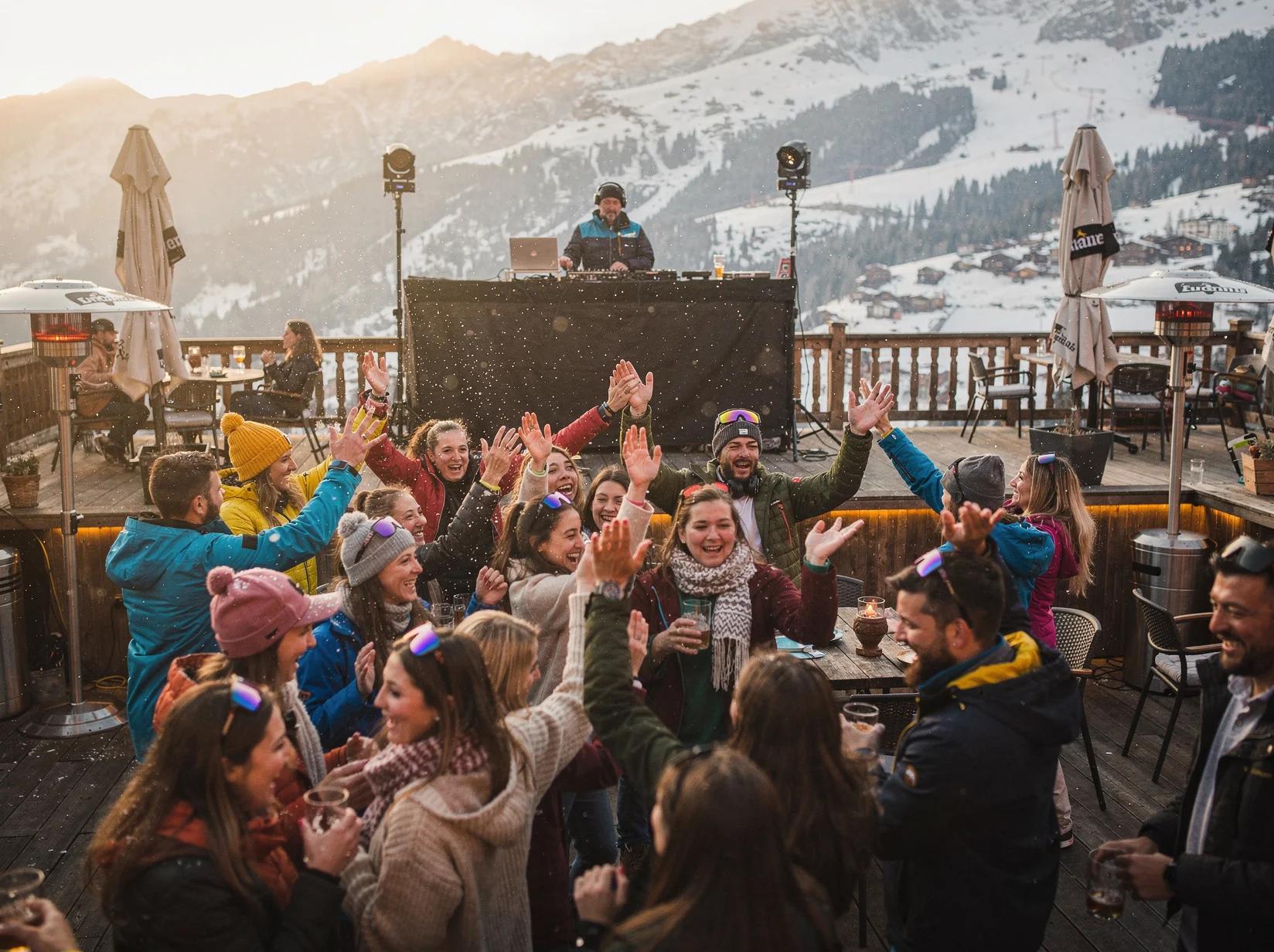 Crowd dancing with arms raised at an apres-ski party while a DJ plays behind the booth