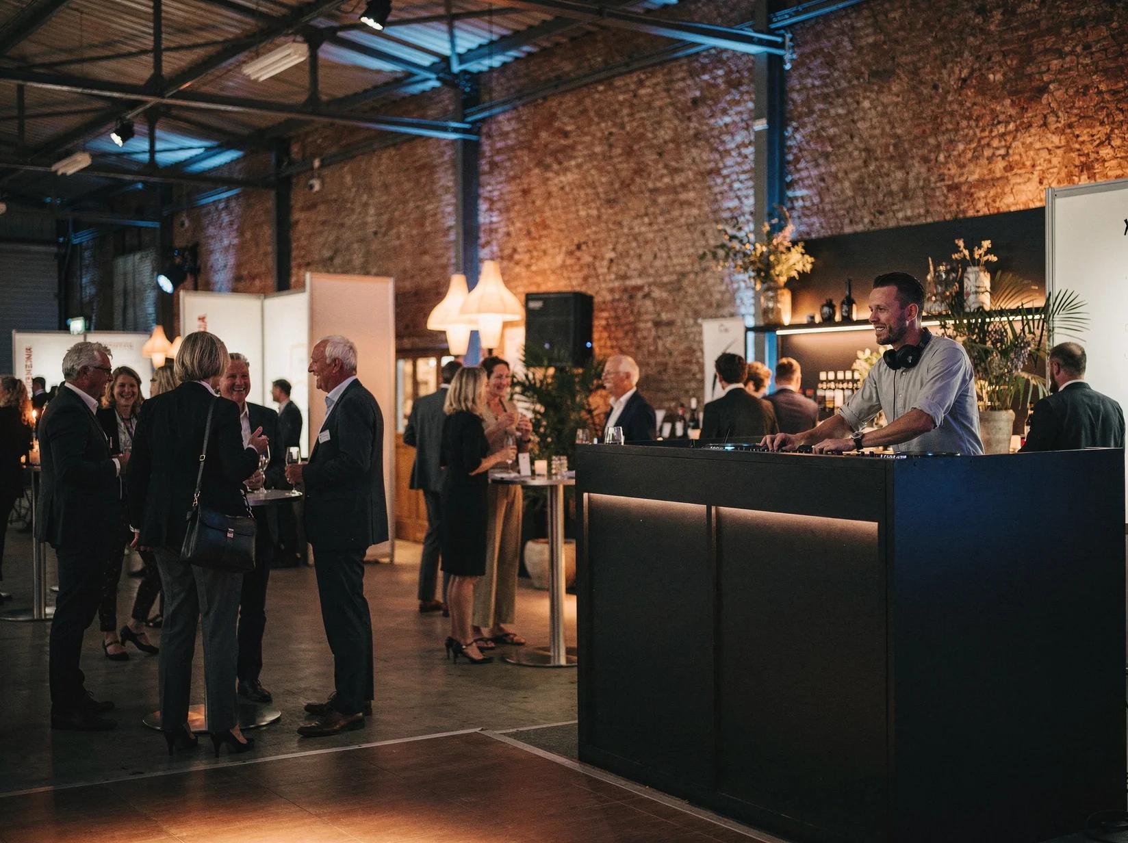 DJ achter een booth tijdens een netwerkevent terwijl professionals in gesprek zijn op de beursvloer