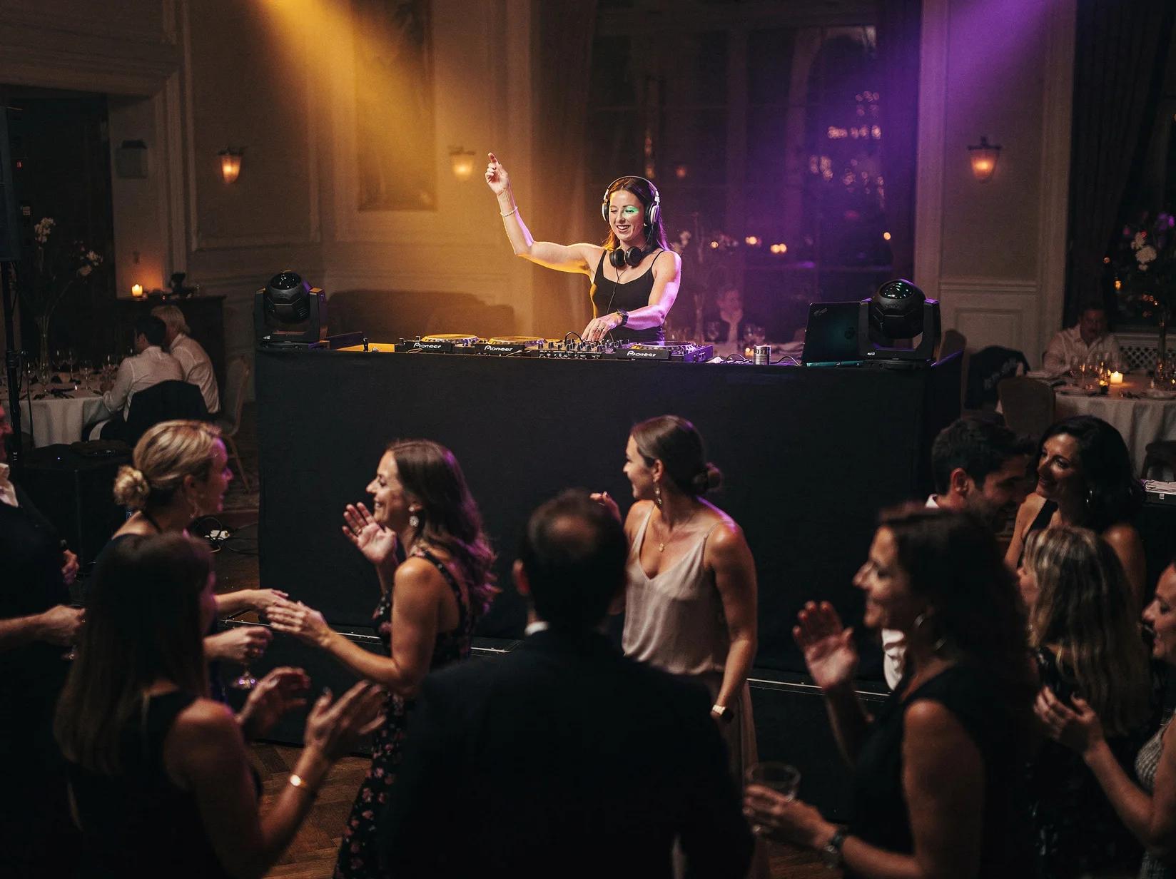 Large crowd dancing at outdoor DJ event with purple and gold stage lighting, string lights, and historic European architecture backdrop at dusk.