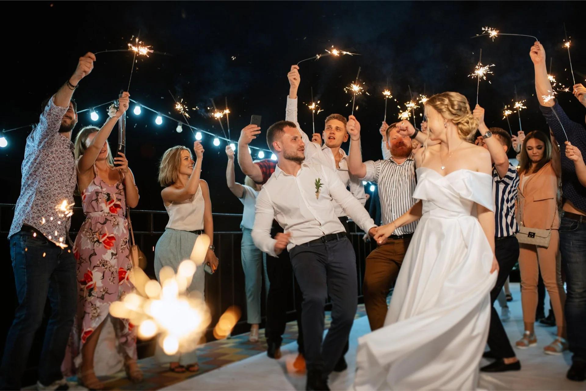Bruidspaar danst met gasten die brandende sparklers omhoog houden tijdens een gezellige avondreceptie met feestverlichting op de achtergrond.