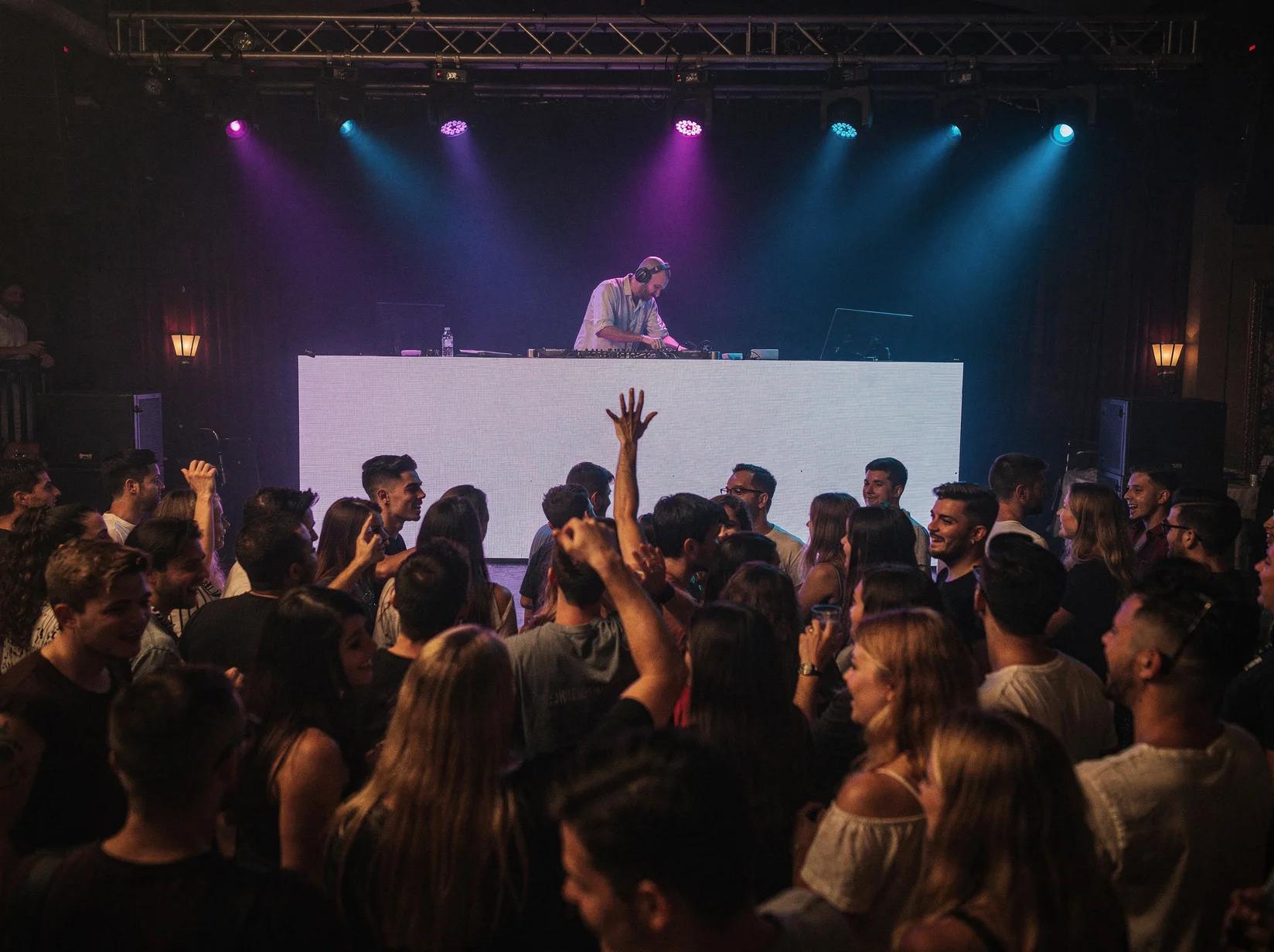 Club DJ draait achter een verlichte booth terwijl een volle dansvloer uitbundig danst