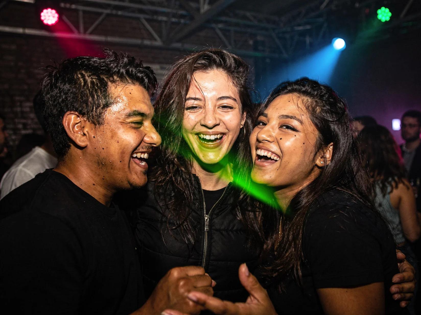 Friends laughing and dancing close together at a drum and bass event lit by warm and neon light
