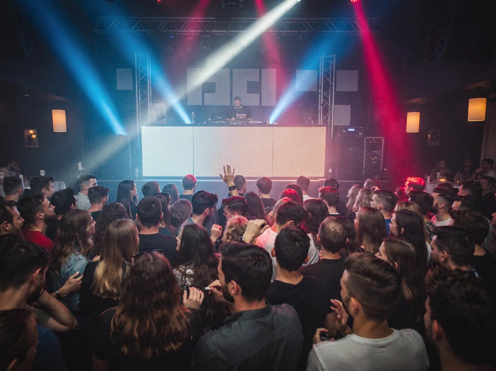 Crowd dancing at full energy in front of a drum and bass DJ in a dark club lit with blue and magenta light