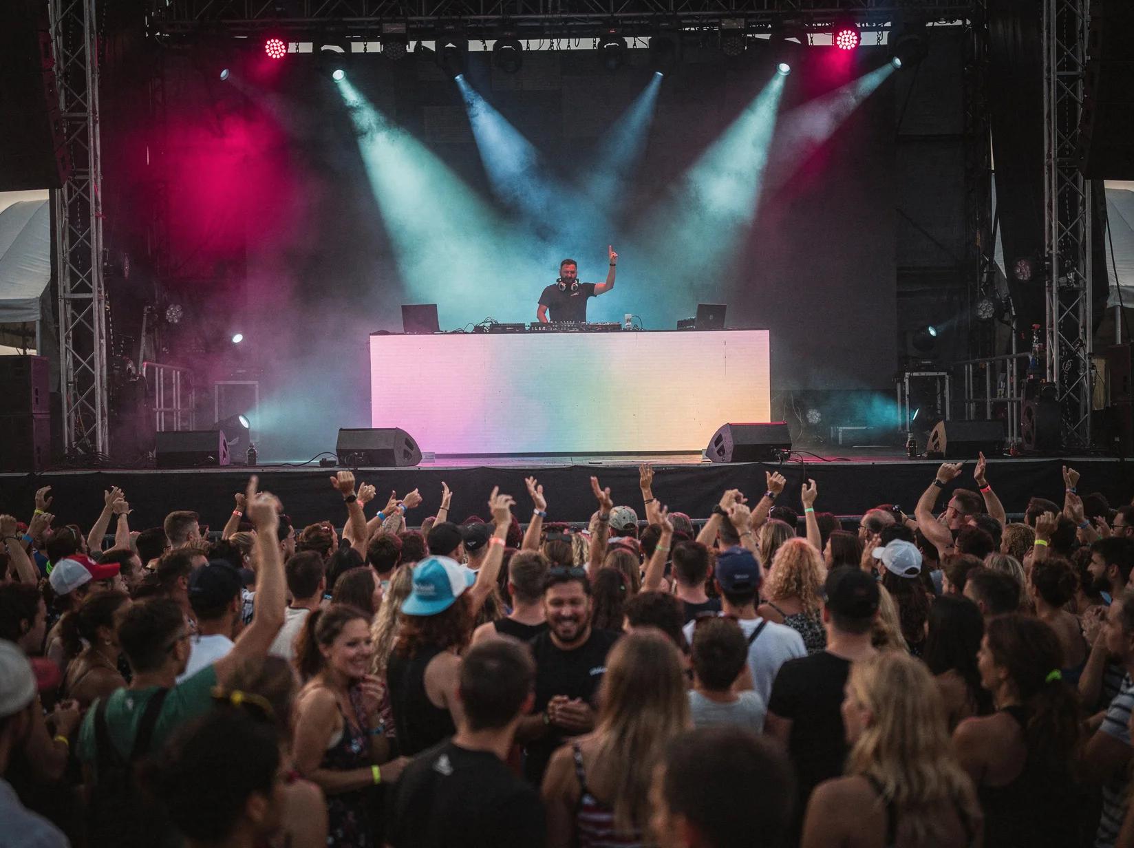 Een festival DJ draait voor een enorme dansende menigte onder kleurrijke podiumlichten op een festivalterrein bij schemer.