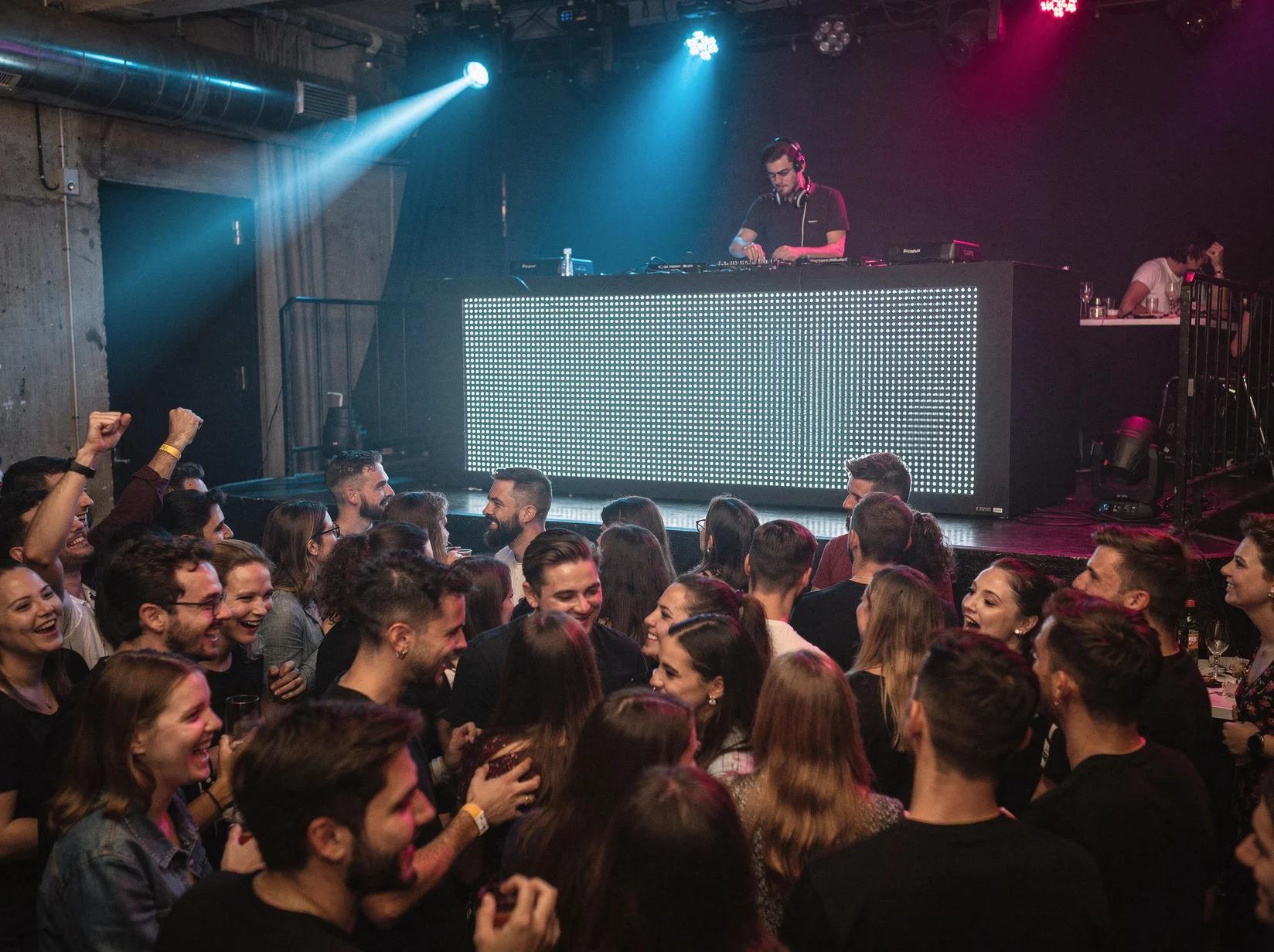 Large crowd with hands raised enjoying a DJ performance at an outdoor European festival during golden hour sunset with city skyline backdrop.