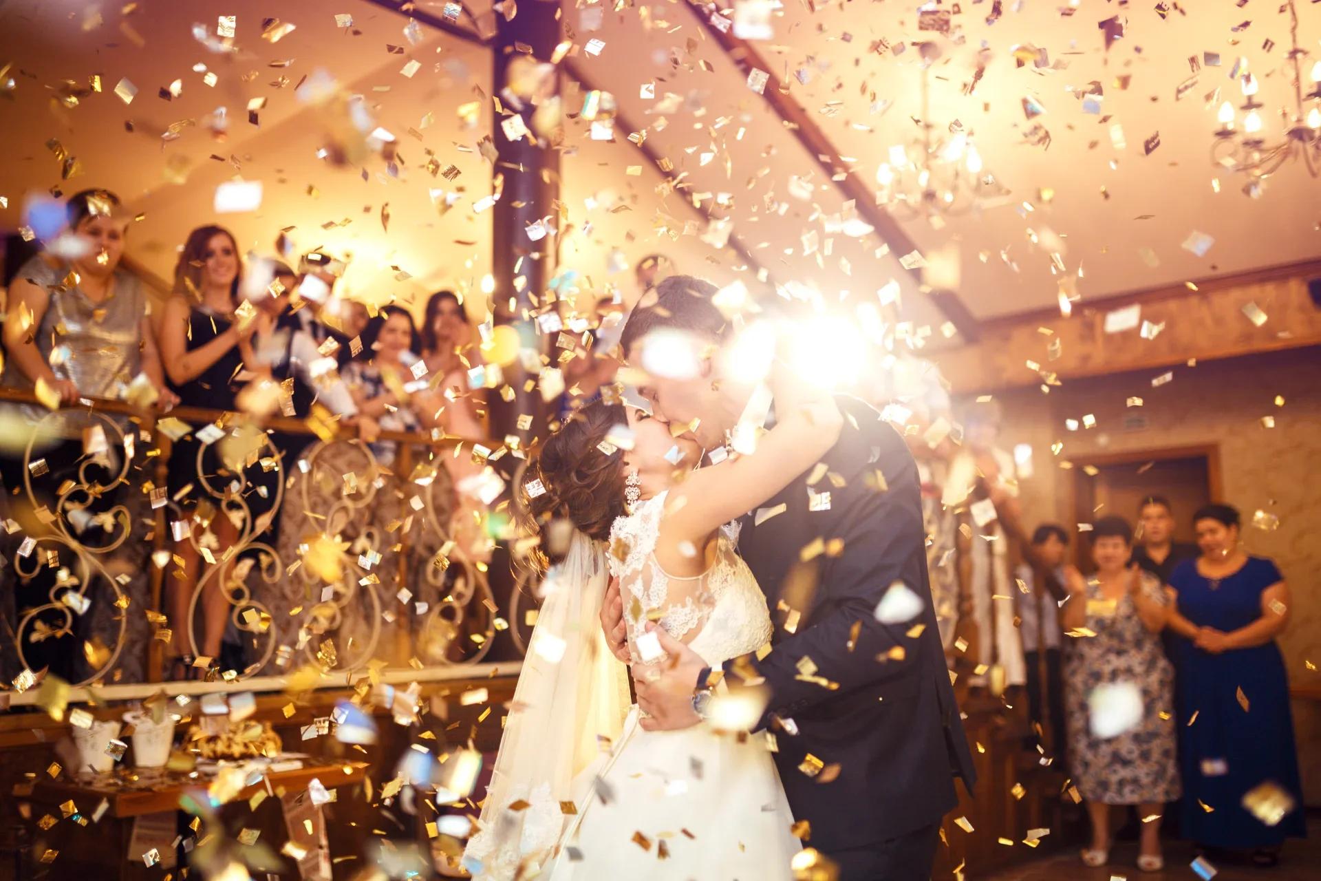 Bruid en bruidegom kussen tijdens hun eerste dans onder gouden confetti terwijl gasten toekijken op een bruiloftsfeest.