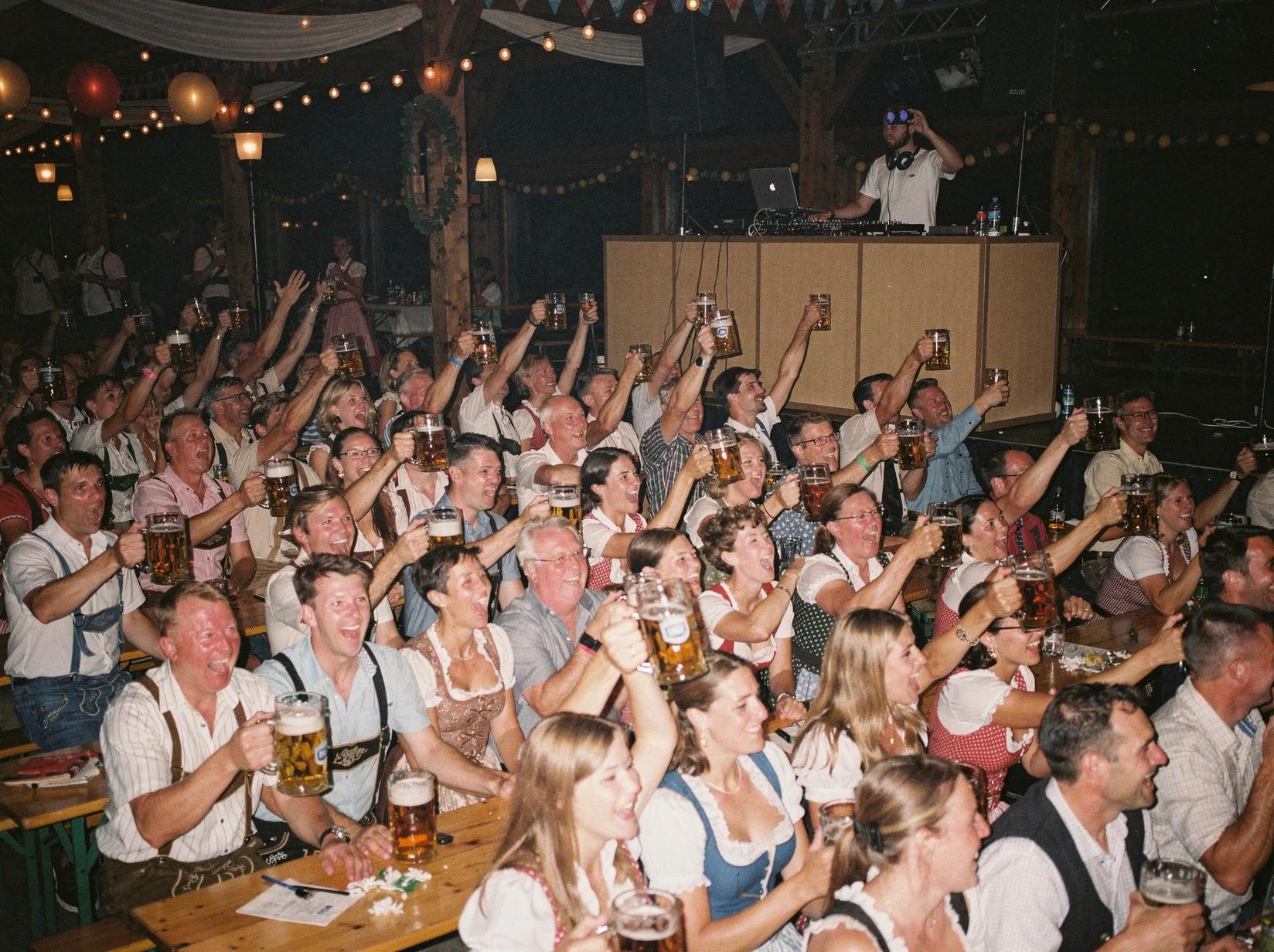 Packed beer tent crowd singing and raising steins while an Oktoberfest DJ plays in the background