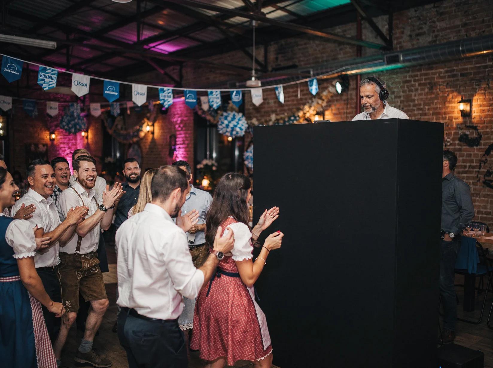Colleagues dancing at an indoor corporate Oktoberfest theme party with a DJ booth in the background