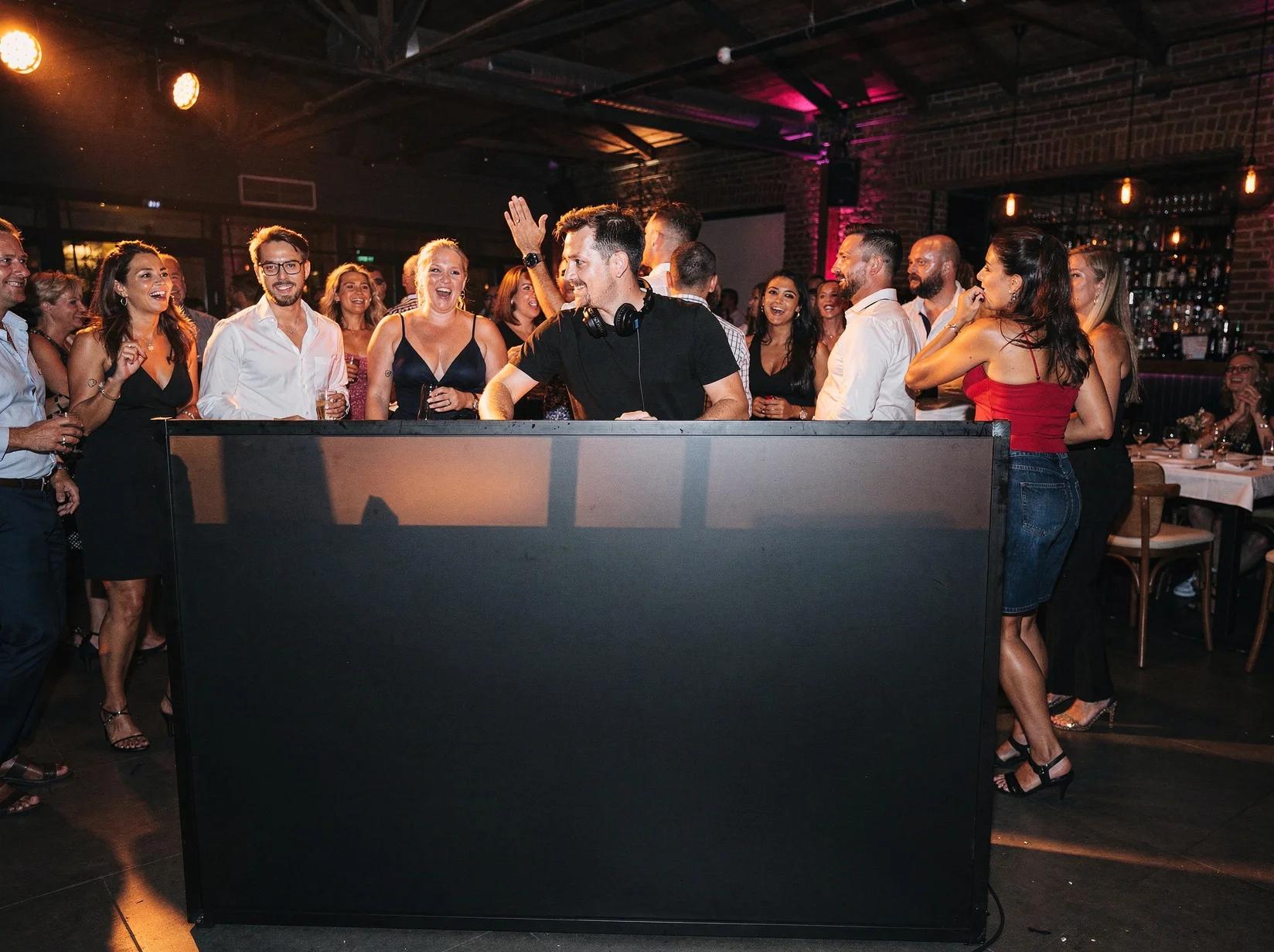 Large crowd dancing and celebrating at an open format DJ party with warm ambient lighting, string lights, and LED backdrop creating vibrant nightlife atmosphere.