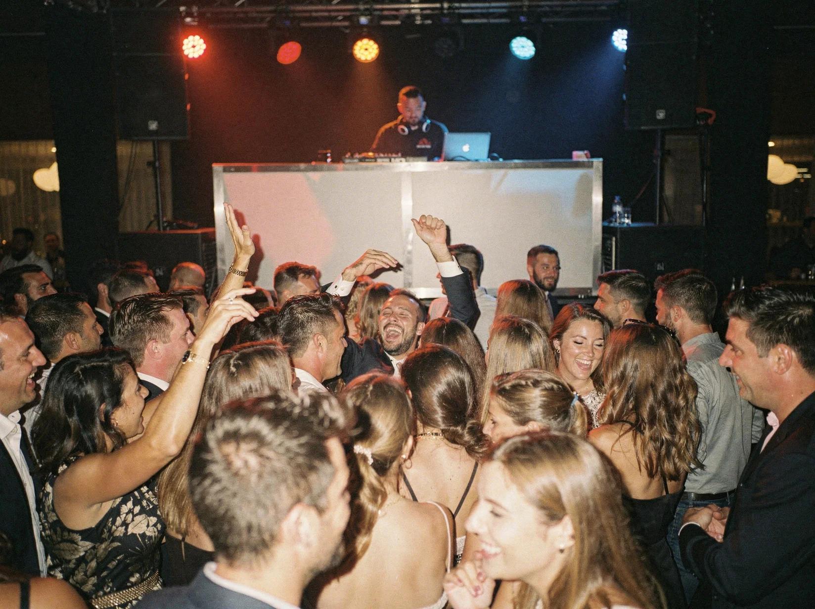 A party DJ fills the dance floor at a packed venue while guests dance with their arms in the air