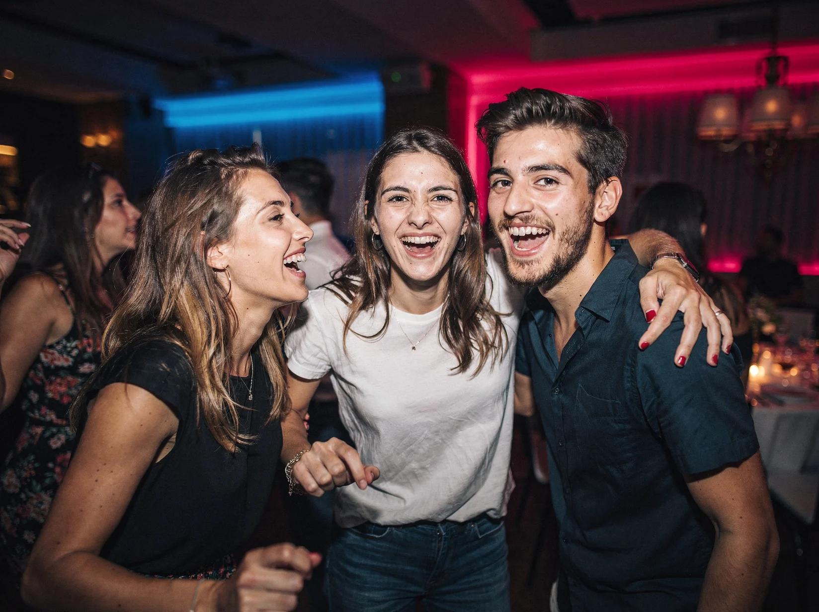 Drie lachende vrienden dansen dicht bij elkaar op een reggaeton-feest met neonkleurig licht