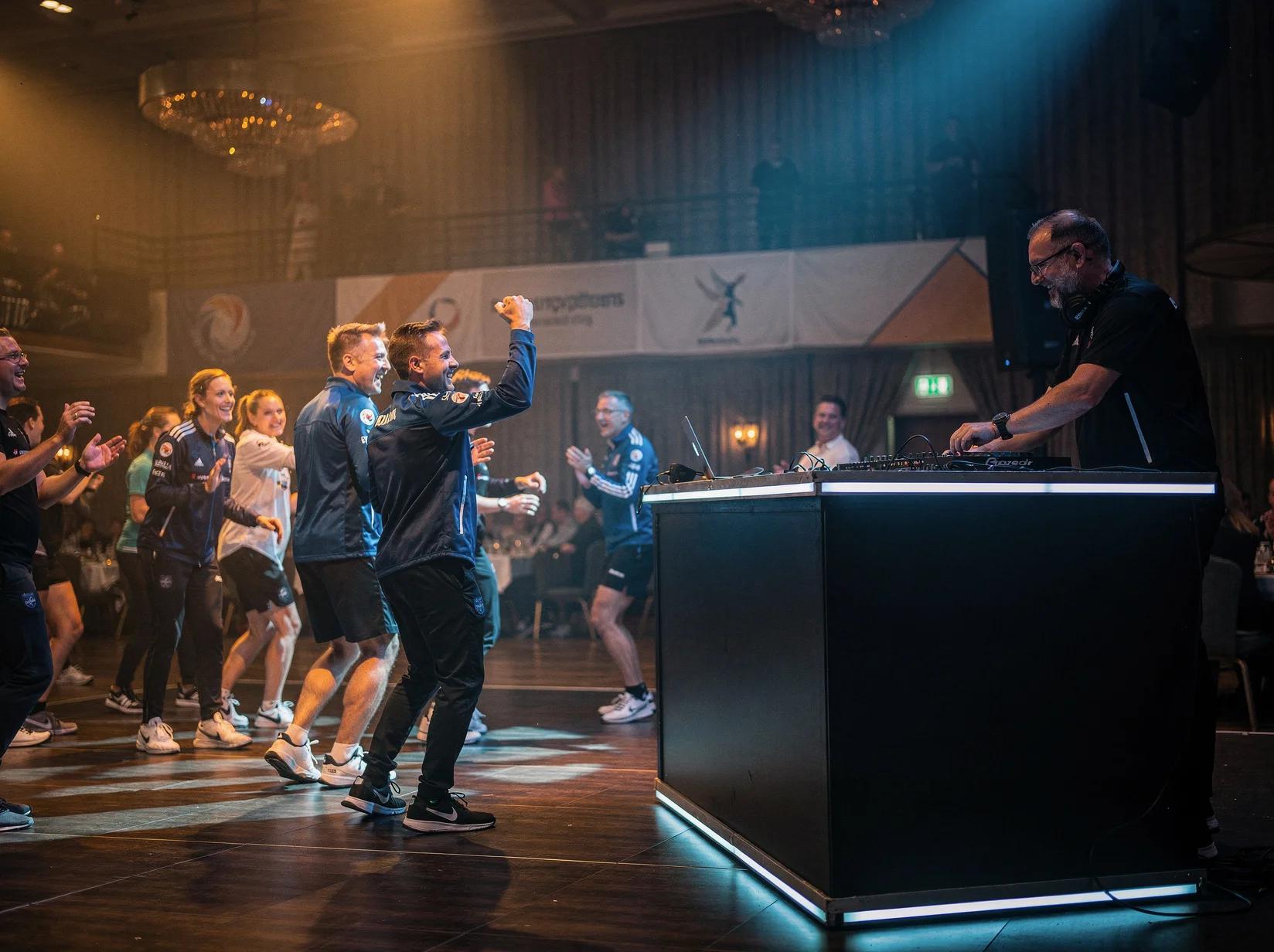 Dansende menigte en DJ tijdens de afterparty van een sportevenement in een sfeervol verlichte sporthal